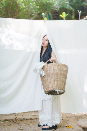A young woman in a flowing white dress stands gracefully outdoors, holding a woven basket under soft sunlight, embodying serenity and joy in nature.の写真素材