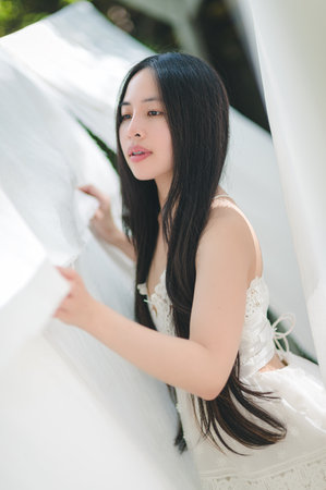 A young woman with long hair poses gracefully amidst flowing white fabric, capturing a moment of serene beauty in soft sunlight. The scene evokes tranquility and elegance.の写真素材