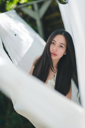 A portrait of a young woman with long black hair immersed in flowing white fabric. The serene atmosphere and soft sunlight create a tranquil and elegant outdoor setting.の写真素材