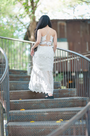 A woman in an elegant white dress walks gracefully up a rustic metal staircase surrounded by nature, showcasing a blend of style and tranquility in an outdoor setting.の写真素材