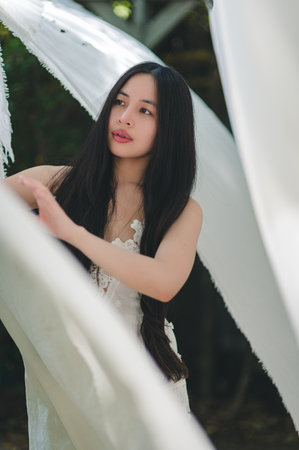 A graceful young woman poses among flowing white fabrics, capturing serene beauty. Natural light enhances her delicate features in this tranquil outdoor setting.の写真素材
