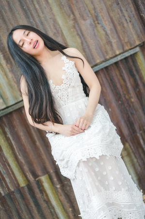 A graceful woman showcases her beauty in a stunning white dress, standing confidently against a rustic backdrop. The natural light enhances the serene atmosphere, capturing elegance and calmness.の写真素材