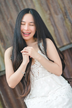 A joyful young woman in a white dress smiles against a rustic backdrop, capturing a moment of pure happiness and charm. Her long hair flows naturally as she expresses feelings of joy and serenity.の写真素材