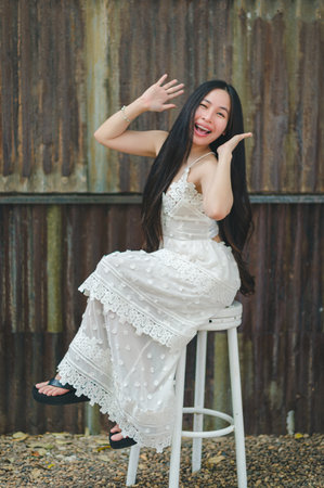 A joyful woman dressed in a flowing white dress poses playfully on a stool, showcasing her long hair and cheerful expression in a rustic outdoor setting.の写真素材