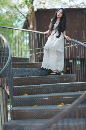 A young woman in a flowing white dress elegantly poses on a spiral staircase surrounded by natural greenery, capturing a serene moment of beauty and tranquility.の写真素材