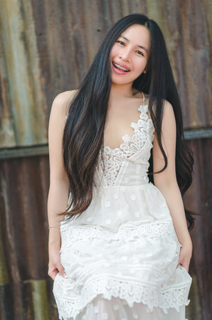A young woman wears a beautiful white dress, smiling warmly while posing outdoors. The soft light enhances her graceful features and long hair.の写真素材