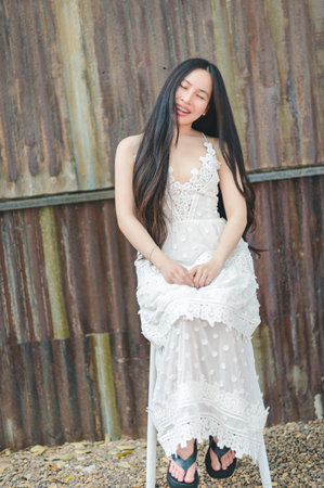 A happy young woman sits gracefully in an elegant white dress against a rustic backdrop, radiating joy and beauty in a serene outdoor setting.の写真素材