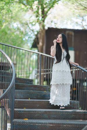 A young woman elegantly poses on a curved metal staircase, surrounded by lush greenery. Her long hair cascades down, complementing the white dress she wears, embodying grace and natural beauty.の写真素材