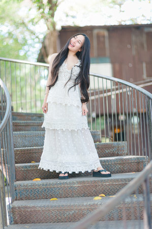 A young woman stands on outdoor stairs, radiating joy in a delicate white dress. The natural setting enhances her cheerful, carefree spirit, creating a serene atmosphere.の写真素材