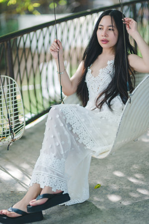 A young woman enjoys a moment of relaxation on a swing, wearing an elegant white dress. The serene outdoor setting captures the essence of summer and tranquility.の写真素材