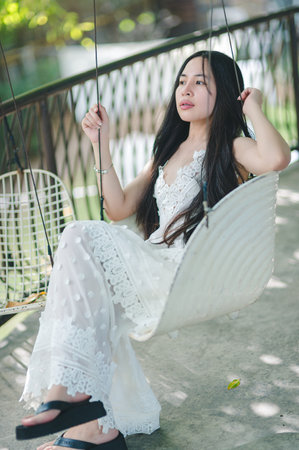 A young woman enjoys a moment of relaxation on a swing surrounded by nature. She exudes calm and elegance in her white dress, capturing a peaceful lifestyle.の写真素材