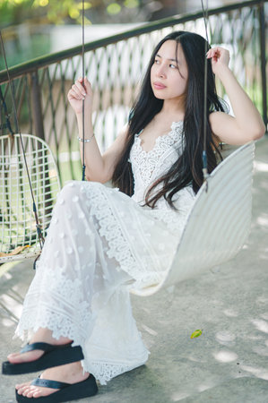 A young woman sits gracefully on a swing, donning a flowing white dress. The serene outdoor setting with natural light creates a peaceful and tranquil atmosphere, perfect for relaxation.の写真素材