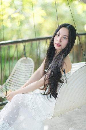 A young woman with long hair elegantly sits on a swing surrounded by lush greenery. She embodies a sense of calm and tranquility in a bright, sunny atmosphere.の写真素材