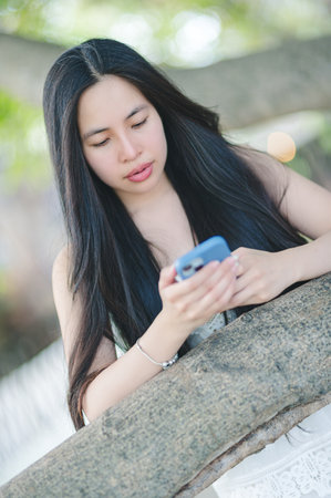A young woman interacts with her smartphone while enjoying a peaceful moment outdoors. Surrounded by nature, she exhibits a relaxed and engaged demeanor.の写真素材
