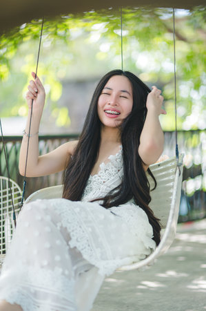 A joyful young woman is swinging under lush greenery, exuding happiness. Her long hair flows as she enjoys a serene moment, embodying carefree leisure.の写真素材