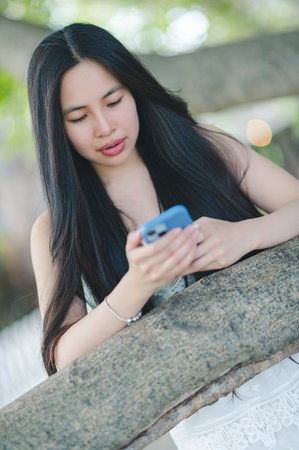 A young woman stands outdoors, engaged with her smartphone while resting her hands on a textured surface, embodying technology and nature in a serene moment.の写真素材
