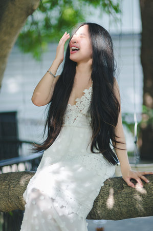 A joyful young woman laughs while sitting on a tree swing. With long hair and a white dress, she embodies carefree happiness in a natural, sunlit setting.の写真素材