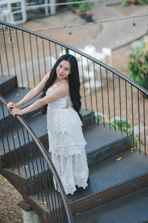 A graceful young woman wearing a white dress poses on a spiral staircase, surrounded by nature. Her smile and relaxed demeanor create a serene atmosphere perfect for lifestyle photography.の写真素材
