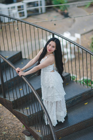 A captivating woman in an elegant white dress poses gracefully on a spiral staircase in an outdoor setting, radiating beauty and confidence.の写真素材