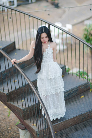 A serene portrait of a woman in a beautiful white lace dress, gracefully ascending winding metal stairs in an outdoor setting. The soft natural light enhances the elegance of the moment.の写真素材