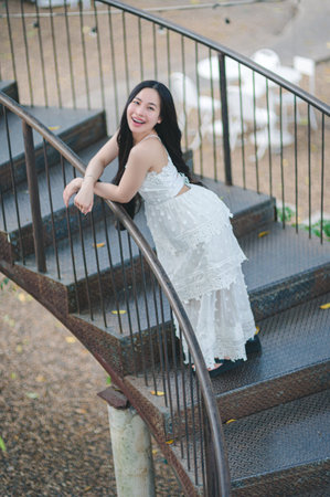 A joyful young woman wearing a white dress poses gracefully on a spiraling staircase, radiating happiness in a serene outdoor environment. She embodies elegance and carefree charm.の写真素材