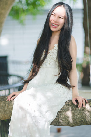 A joyful young woman with long hair enjoys the outdoors while sitting on a tree branch. Her smile radiates happiness, embodying a carefree summer spirit.の写真素材