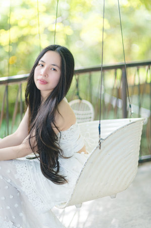 A young woman with long hair sits gracefully on a swing, surrounded by natural light and greenery, embodying serenity and beauty in a peaceful outdoor setting.の写真素材