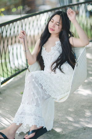 A young woman relaxes on a swing in a serene outdoor setting. She wears a beautiful white dress, embodying tranquility and style. Perfect for lifestyle themes.の写真素材