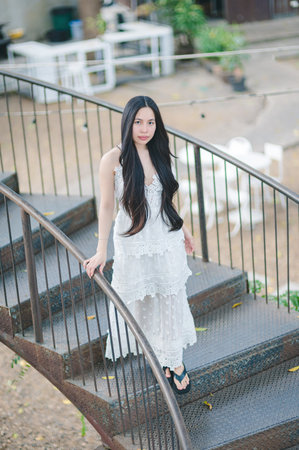 A beautiful woman with long hair wears a flowing white dress while standing gracefully on an outdoor staircase. The serene setting exudes elegance and tranquility.の写真素材