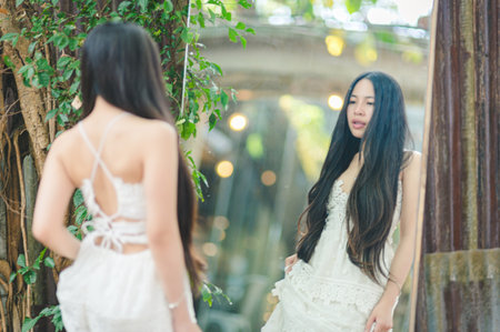 A young woman stands in front of a mirror, gracefully gazing at her reflection. She wears an elegant dress, surrounded by lush greenery, creating a serene atmosphere.の写真素材