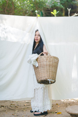 A young woman stands gracefully in a white dress, holding a woven basket amidst hanging fabrics in a tranquil outdoor setting, embodying serenity and beauty.の写真素材