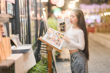 A young woman enjoys her drink while studying a menu on a lively city street. The warm ambiance and colorful lights create a cozy atmosphere perfect for dining.の写真素材