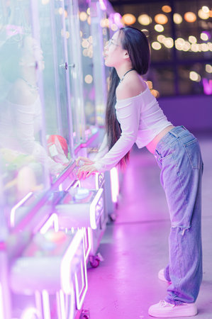 A young woman dressed in a casual outfit leans over an illuminated arcade game machine, reflecting the vibrant neon lights. The playful atmosphere captures a moment of joy and excitement in a nightlife setting.の写真素材