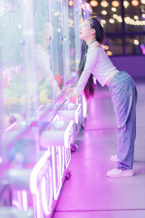 A girl interacts with a claw machine in an atmospheric amusement arcade. Neon lights create a vibrant backdrop, highlighting her playful spirit and reflection.の写真素材