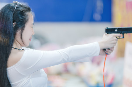 A young woman playfully aims a toy gun, smiling in a vibrant indoor setting. The scene captures joy and youthful excitement, highlighted by colorful surroundings.の写真素材