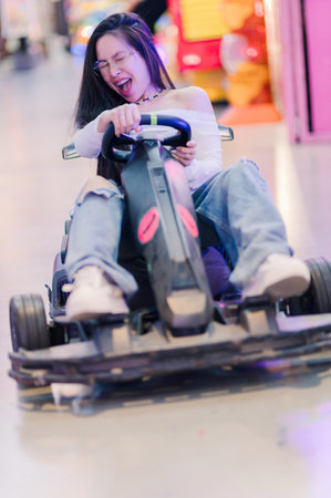 A young woman showcases her thrill and excitement as she joyfully rides a go-kart in a vibrant amusement arcade, surrounded by colorful lights and lively atmosphere.の写真素材