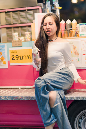A young woman sits casually enjoying ice cream from a vibrant food truck, embodying summer joy and trendy style in a lively urban environment.の写真素材