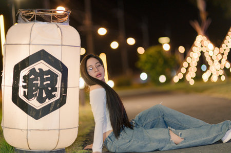 A young woman poses gracefully beside a glowing lantern at night. Her long hair flows as she enjoys the serene atmosphere, creating a beautiful and captivating scene.の写真素材