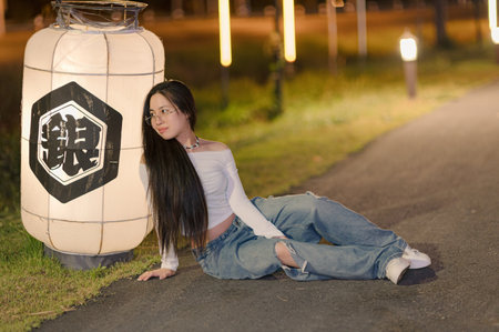 A young woman elegantly poses by a glowing lantern in a tranquil outdoor setting at night, showcasing a blend of fashion and serene ambiance.の写真素材