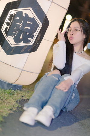 A young woman poses playfully beside a glowing lantern, capturing a vibrant nighttime street scene. Her casual outfit and joyful expression create a lively atmosphere.の写真素材