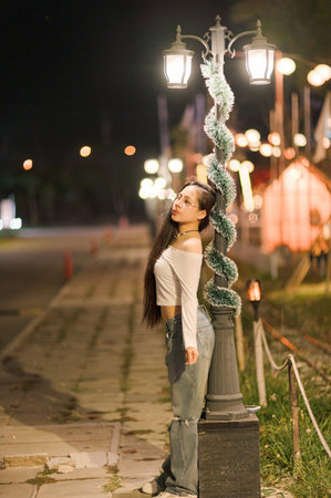A young woman with long hair poses elegantly by a decorative street lamp at night. The scene captures urban charm and a stylish vibe in a serene atmosphere.の写真素材