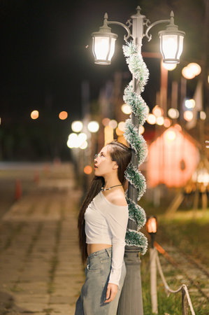 A young woman stands by a street lamp, gazing upward in a serene urban environment. The night setting is enhanced by decorative lights and a festive atmosphere.の写真素材