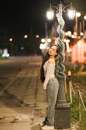 A young woman stands elegantly by a street lamp at night, showcasing a stylish urban look. The soft glow from surrounding lights creates a serene atmosphere, perfect for capturing beauty and tranquility in an outdoor setting.の写真素材