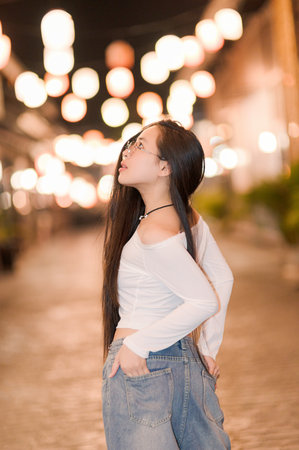 A captivating portrait of a woman with long hair standing in a charming city alley at night, surrounded by soft bokeh lights, exuding a serene and thoughtful mood.の写真素材
