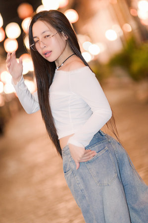 A young woman poses gracefully outdoors at night, showcasing her long hair and chic style amidst blurred city lights, capturing a moment of urban elegance.の写真素材