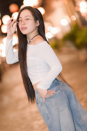 A stylish young woman poses confidently outdoors, surrounded by warm city lights. Her casual yet trendy outfit and long hair create a captivating and relaxed vibe.の写真素材