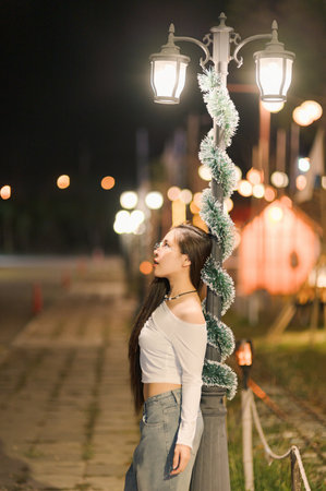 A young woman poses beside a charming street lamp at night, showcasing an elegant urban scene. The gentle glow of the lamp highlights her relaxed expression and casual style.の写真素材
