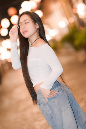 A young woman poses confidently in a casual outfit on a vibrant street, illuminated by soft bokeh lights. Her long hair and trendy style reflect a modern, relaxed lifestyle.の写真素材