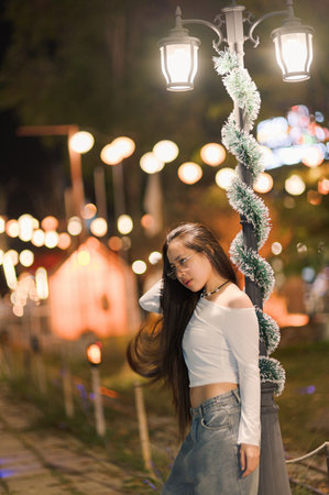 A young woman poses gracefully by a decorative street lantern in a festive evening setting, showcasing a vibrant atmosphere filled with glowing lights and joy.の写真素材
