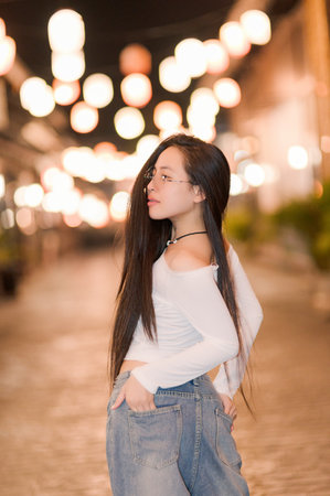 A stylish young woman poses eloquently in a charming urban setting, showcasing her long hair against a backdrop of glowing lanterns, capturing a serene night ambiance.の写真素材